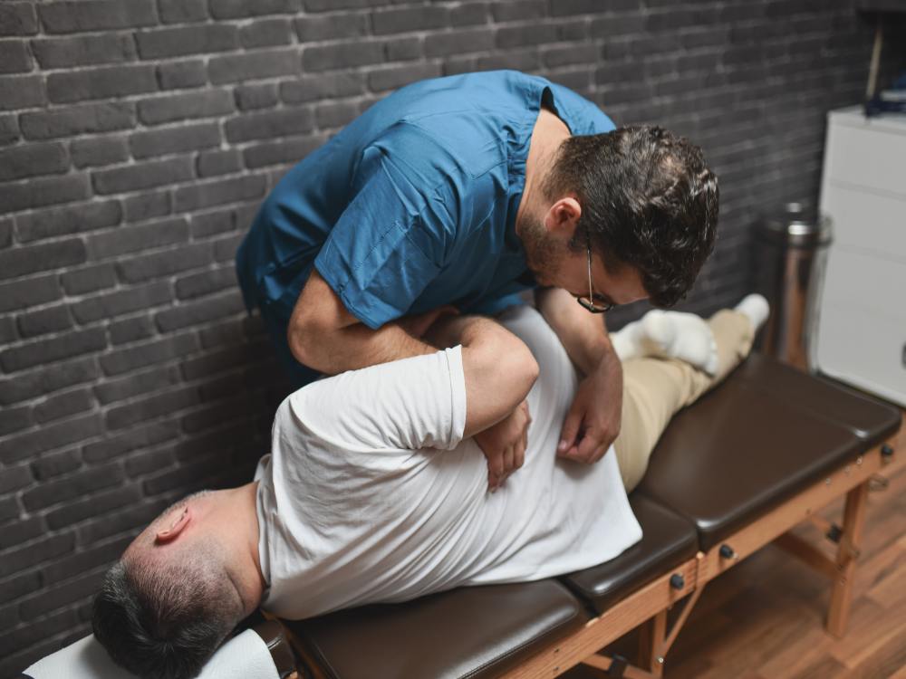 A male chiropractor applies a chiropractic adjustment to the back of a male patient who is laying on his side on an exam table.